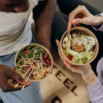 Two people holding colorful bowls filled with leafy greens, whole-grain pasta, beans, and fresh vegetables—illustrating nutrient-dense eating habits that support healthy aging and longevity.