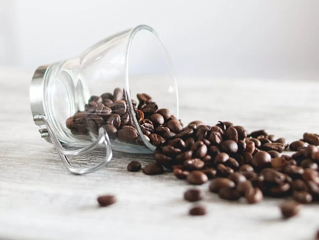 Glass jar tipped over with roasted coffee beans spilling out onto a light wooden surface—illustrating the question of whether coffee contributes to aging or supports longevity.
