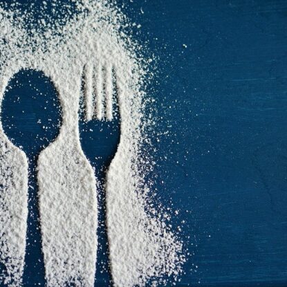 Artistic silhouette of a spoon and fork outlined in scattered white powder on a dark blue background, symbolizing dietary choices and their role in promoting slow aging through mindful eating.