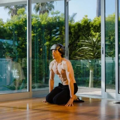 A shirtless man wearing a brain-monitoring helmet kneels on the floor in a bright, modern room, embodying the intense biohacking routine of the Blueprint Project aimed at reversing aging through technology, exercise, and data-driven self-optimization.