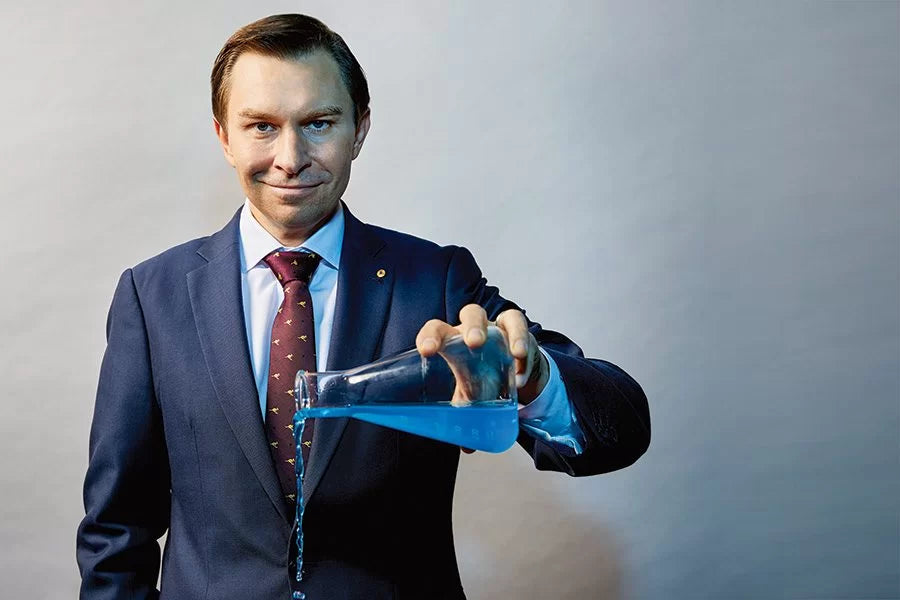 Dr. David Sinclair, Harvard geneticist and longevity researcher, wearing a navy suit and pouring blue liquid from a lab flask—symbolizing his pioneering work in anti-aging science and NAD+ research.