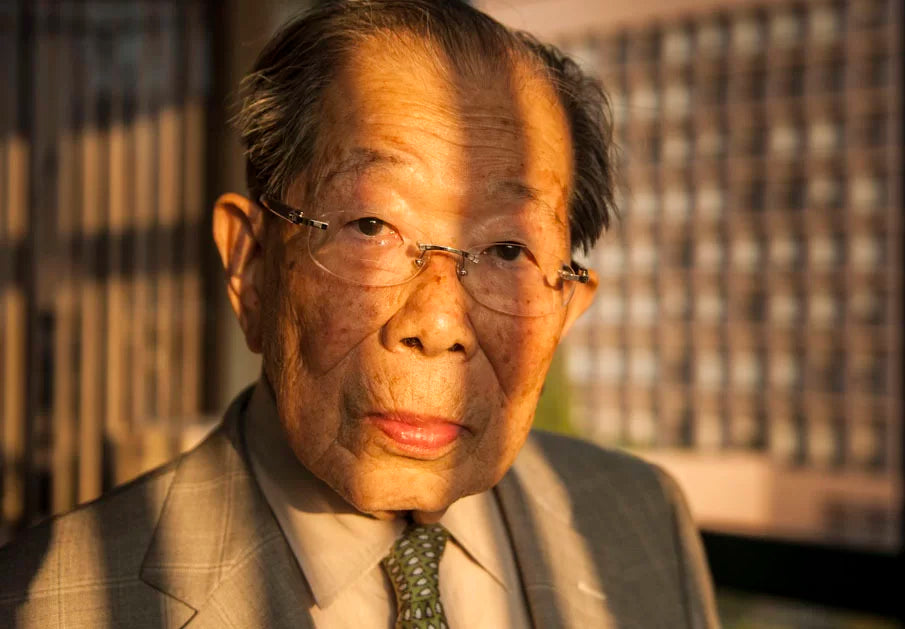 Portrait of Dr. Shigeaki Hinohara, a renowned Japanese physician and longevity expert, wearing a suit and glasses in warm evening light.