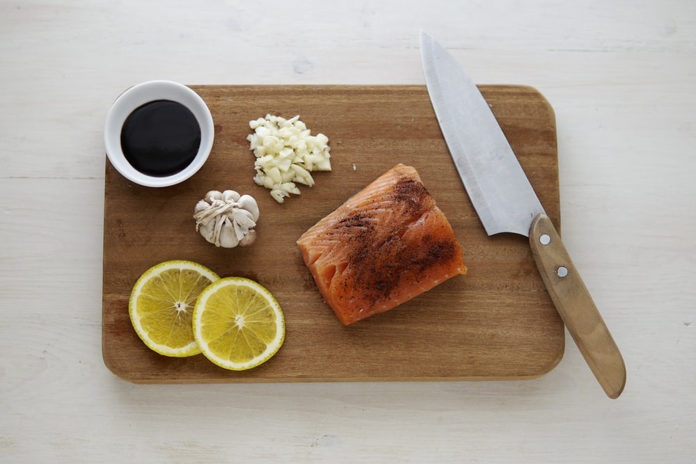 Soy sauce, garlic, lemon, a piece of salmon, a knife on a cutting board on a white table.